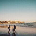A family enjoying a peaceful walk along Bondi Beach during sunset, with city views in the background.