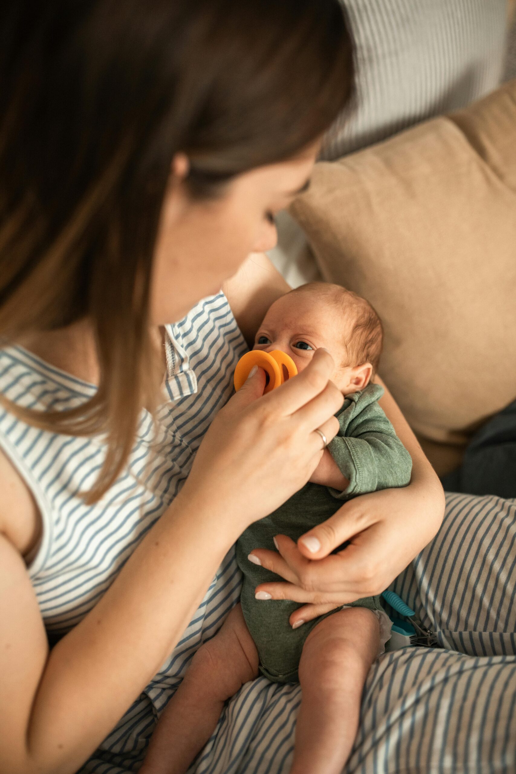 A mother tenderly feeds her newborn baby with a bright orange pacifier indoors.